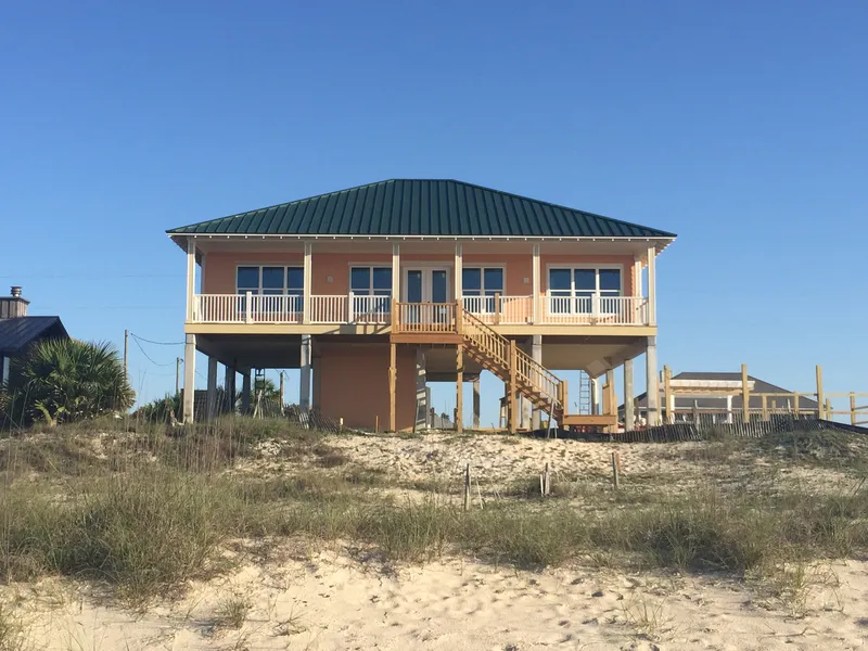 Coastal home with a green hip roof and wide porches on pilings above the dunes
