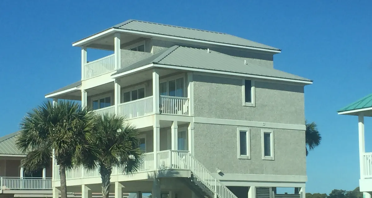 Three-story elevated coastal home with a sage-green metal roof, stacked porches and palm trees