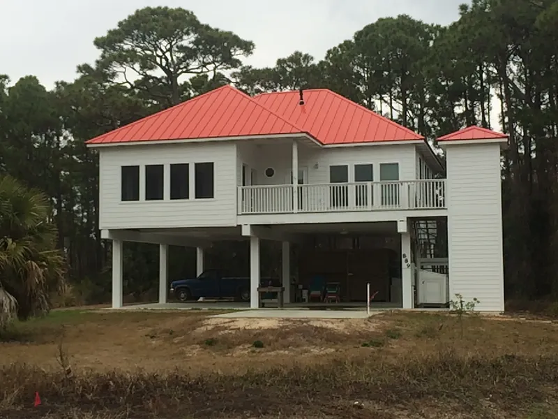 White elevated home with a red metal roof and covered parking underneath, set among pines