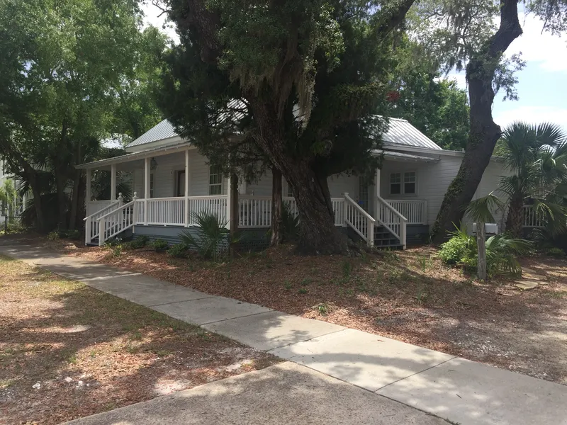 Low white cottage with a porch railing under large live oaks